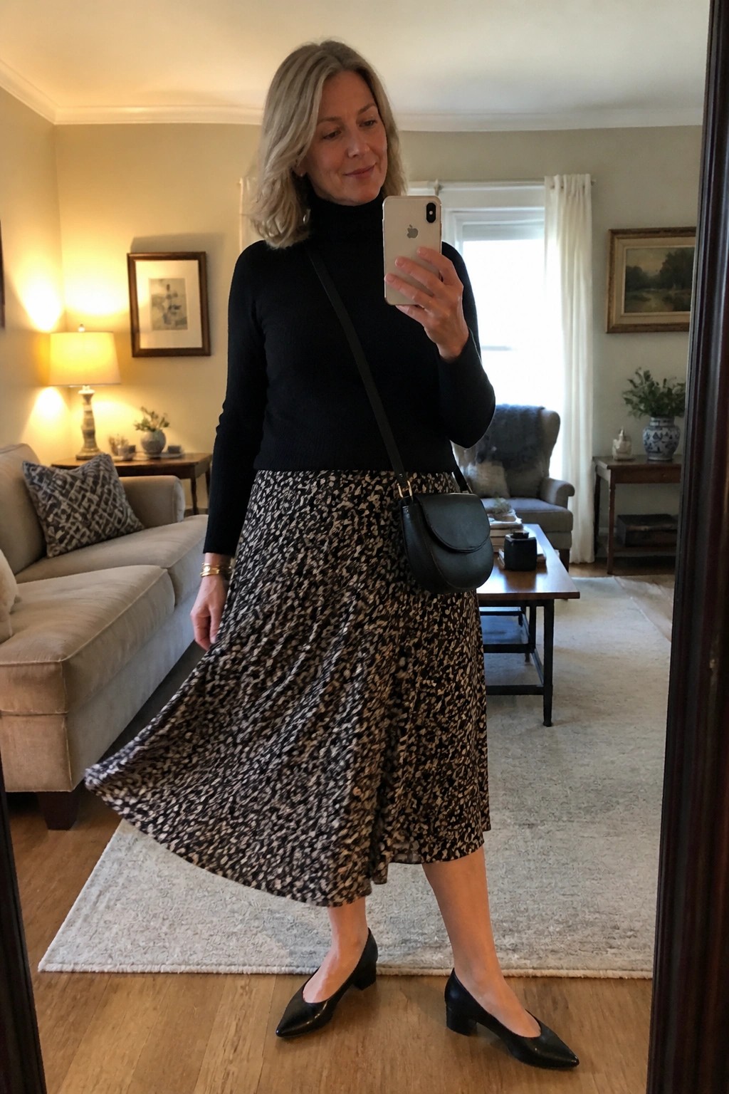 Woman in black turtleneck and patterned skirt taking mirror selfie in living room