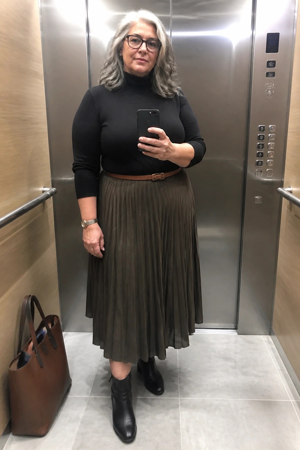Gray-haired woman in black turtleneck, olive pleated skirt, and boots selfies in elevator