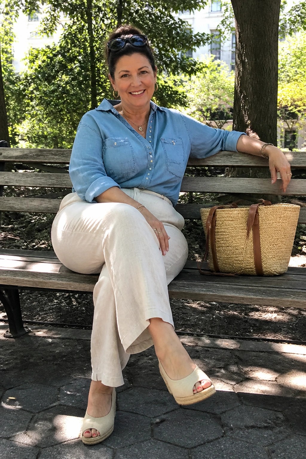 Woman over 50 wearing a light blue chambray shirt tucked into cream wide leg linen pants with beige espadrille wedges and a straw tote bag