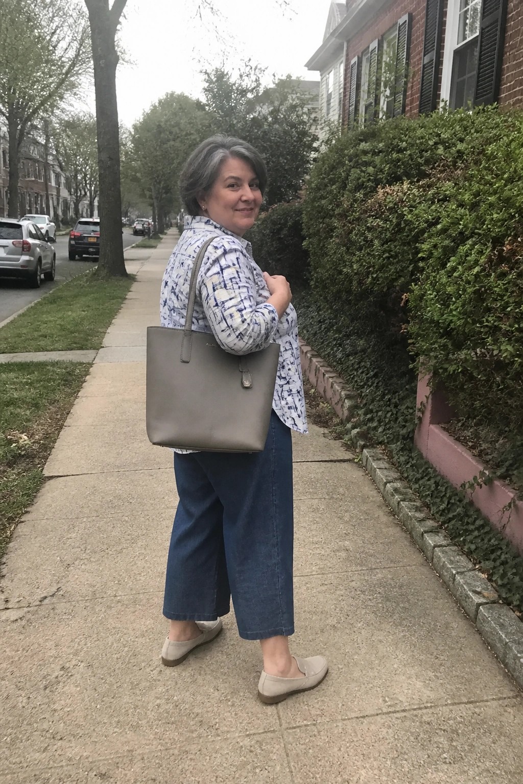 Woman over 50 wearing a blue and white abstract print shirt tucked into wide leg blue denim culottes with beige loafers and carrying a gray tote bag on a sidewalk