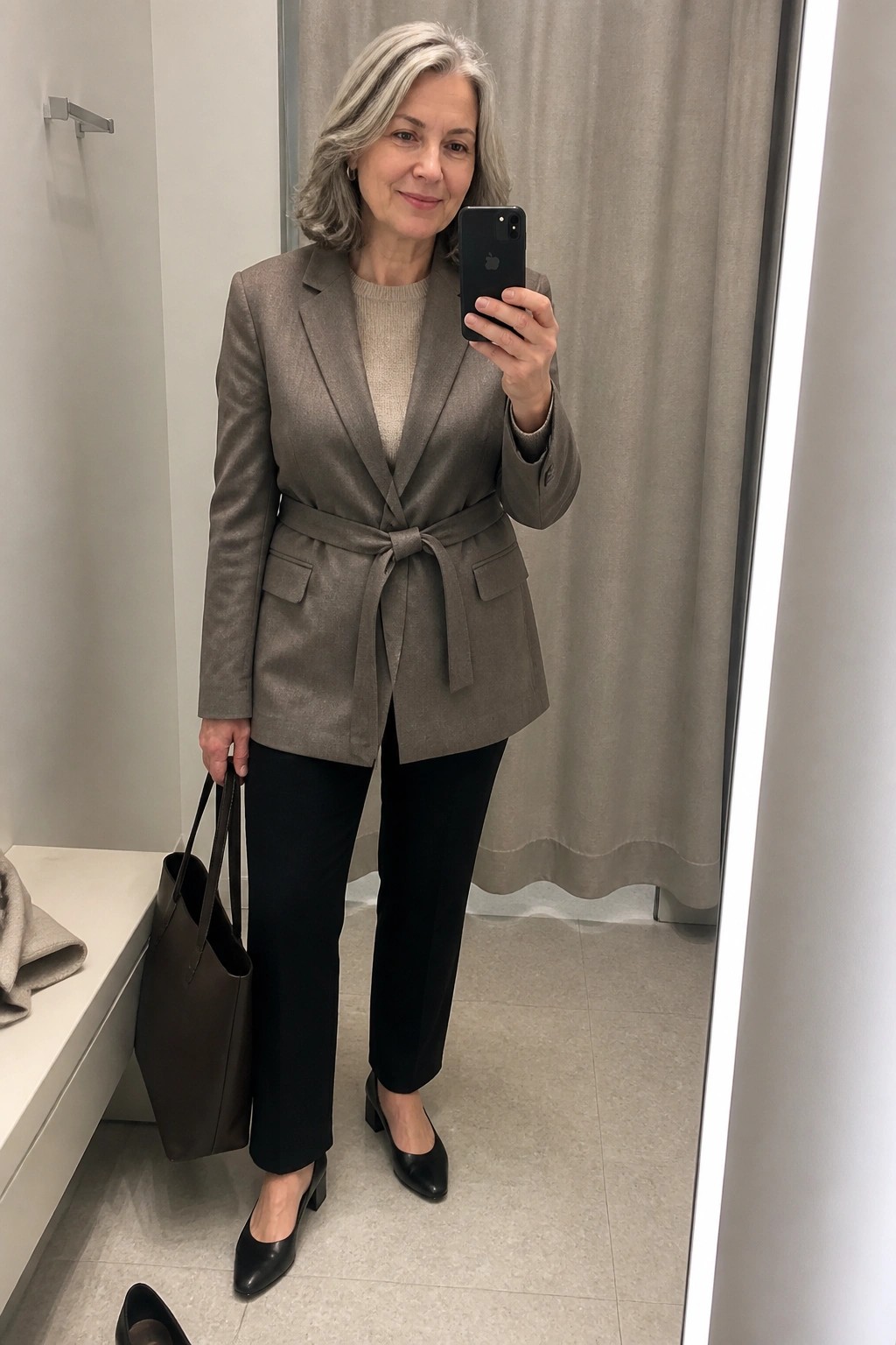 Woman wearing a taupe belted blazer over a beige knit sweater paired with black pants and black block heel shoes while holding a brown leather tote bag in a fitting room mirror selfie