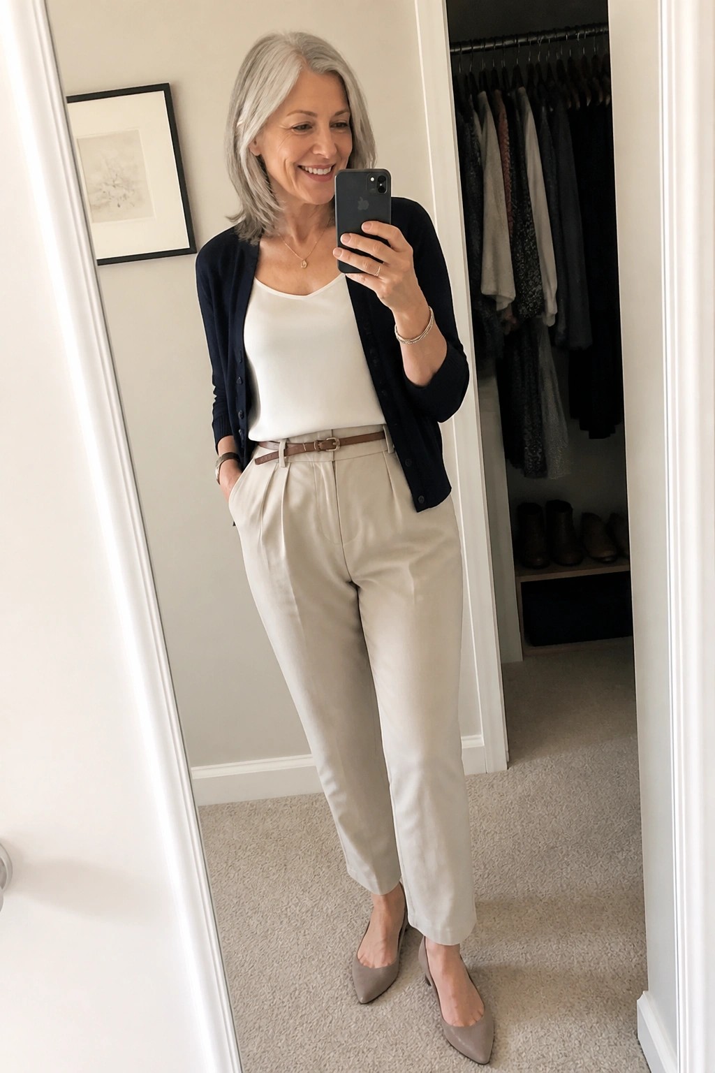 Smiling gray-haired woman in navy cardigan and beige trousers taking mirror selfie.