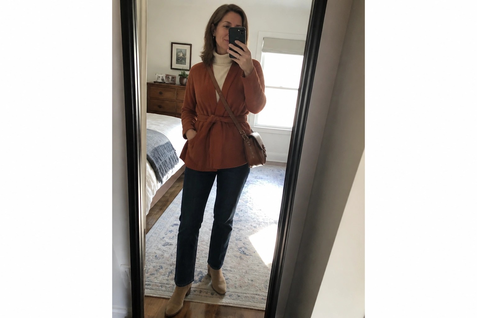Woman wearing a rust belted cardigan over a cream turtleneck with blue jeans and tan suede boots while holding a phone for a mirror selfie.