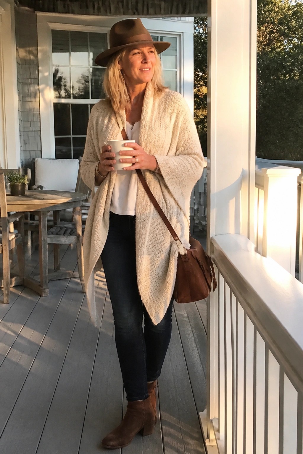 Blonde woman in cream cardigan and brown hat holds mug on sunny porch.