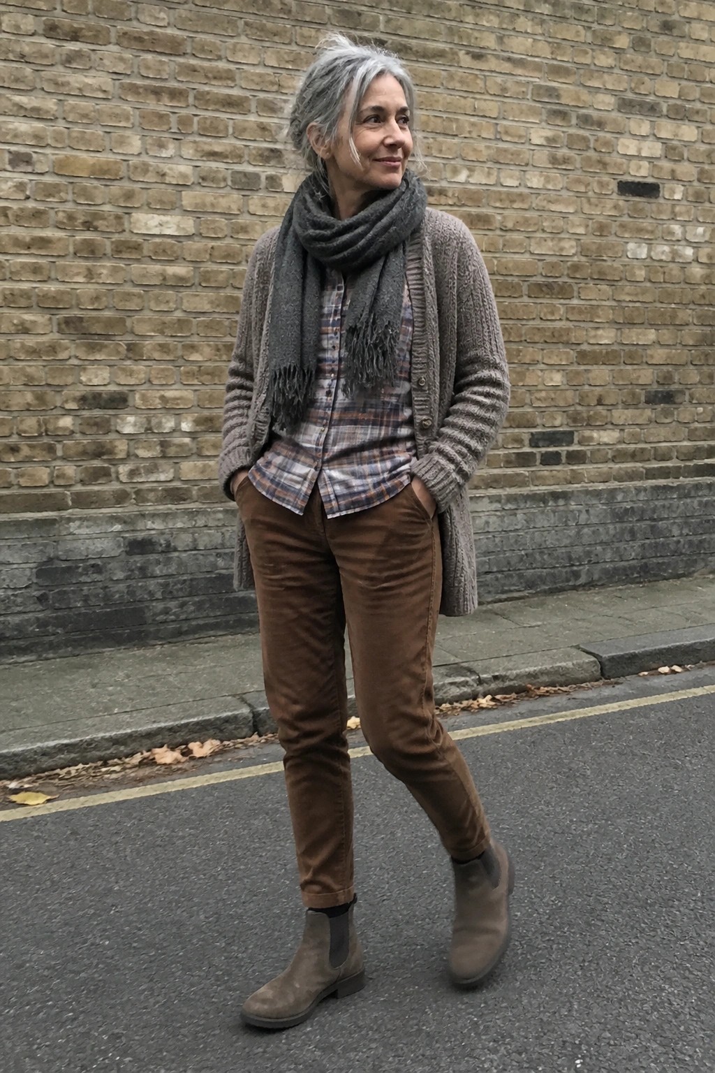 Woman with gray hair in plaid shirt, brown pants, gray scarf on urban street.