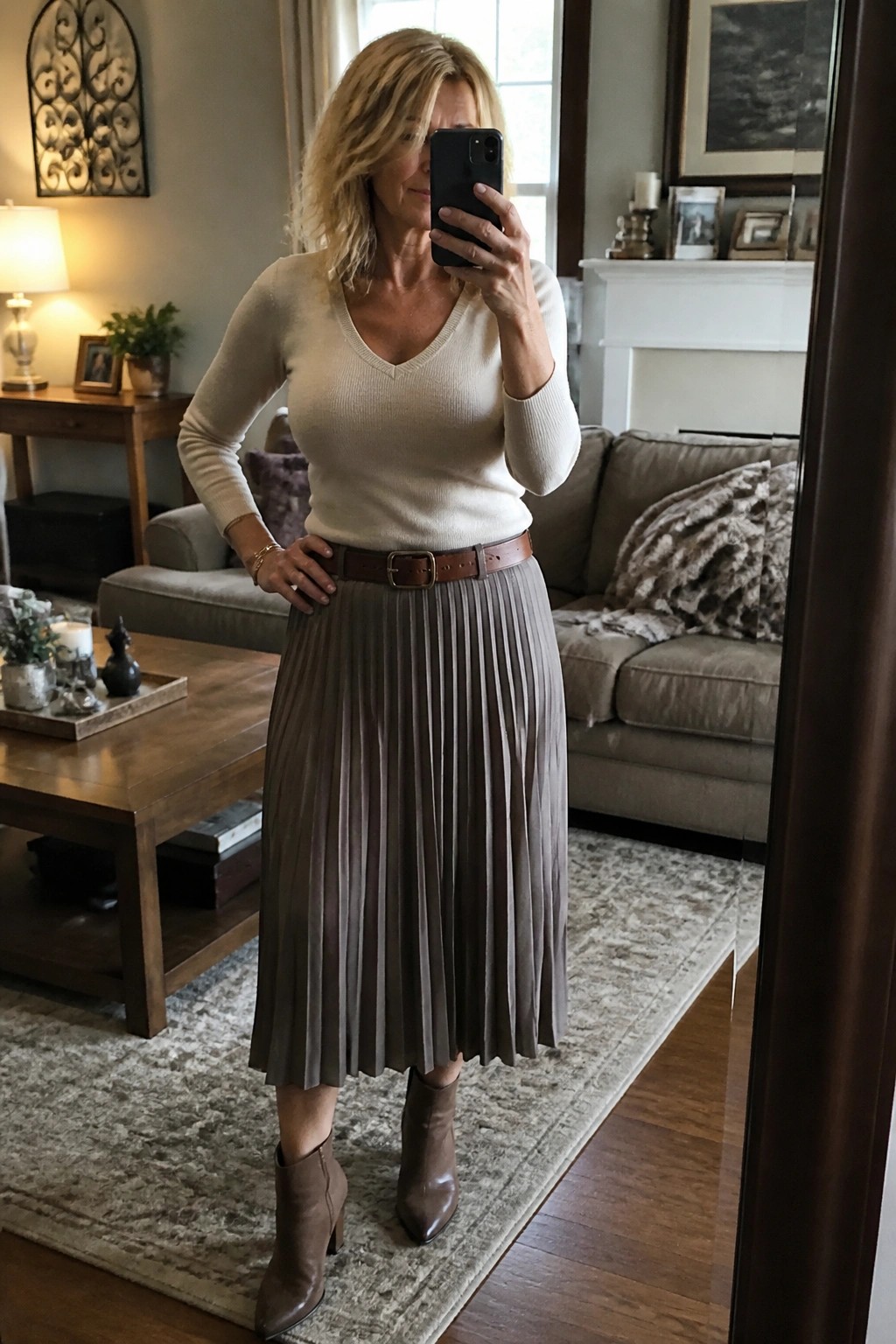 Woman in cream sweater and taupe pleated skirt taking mirror selfie in living room