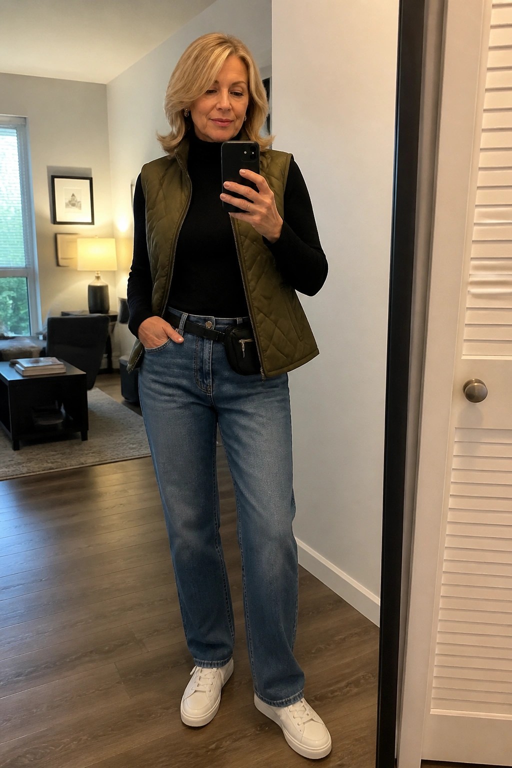 Blonde woman in olive quilted vest, black turtleneck, blue jeans, and white sneakers taking mirror selfie.