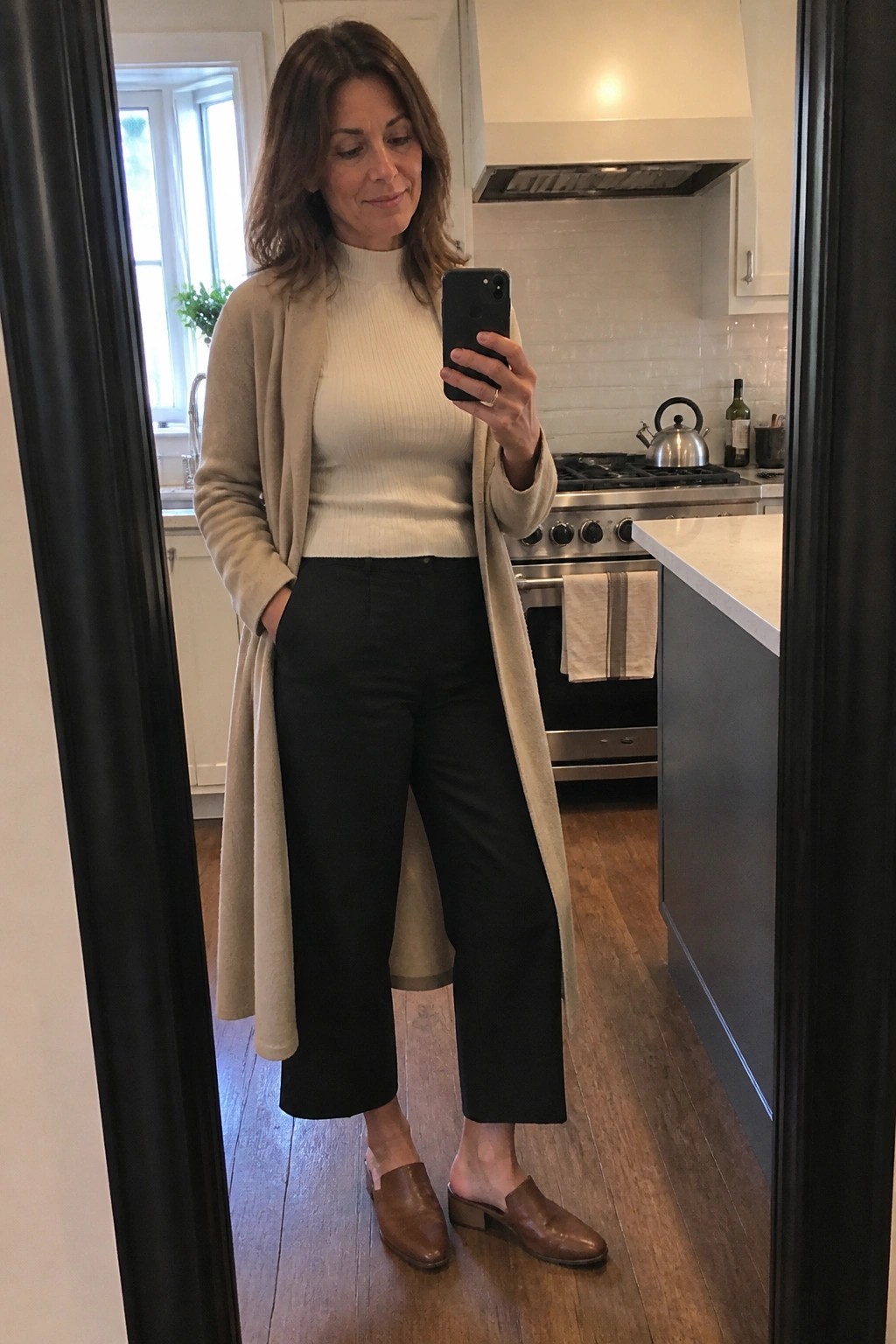 Woman in beige cardigan and black pants takes mirror selfie in kitchen.