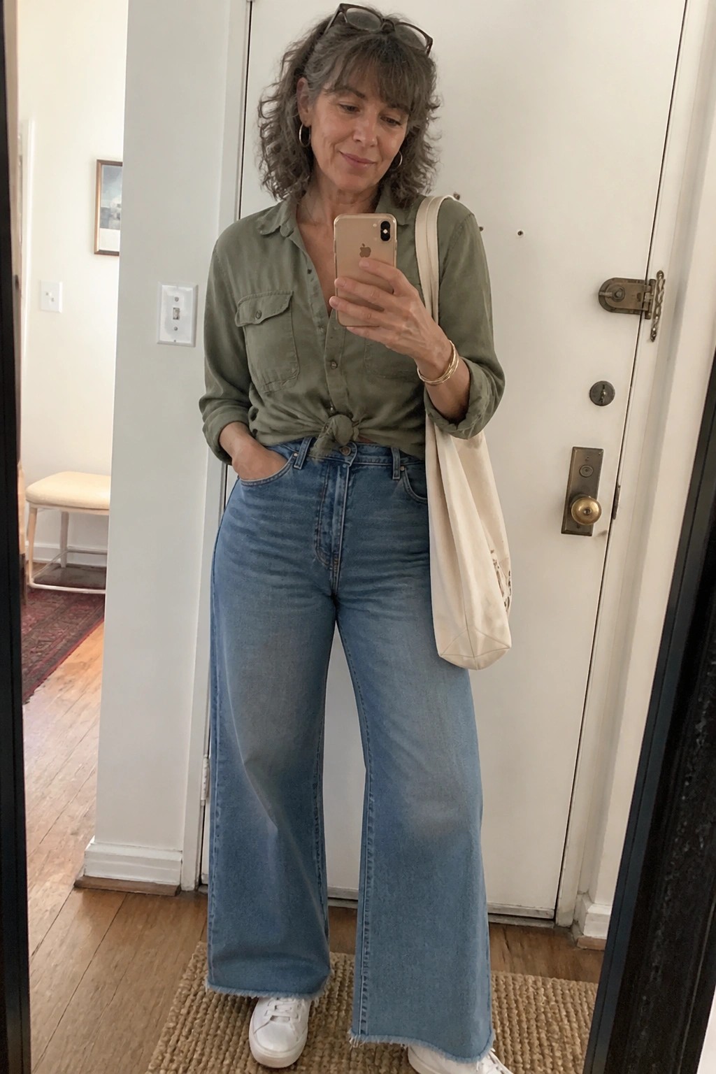 Woman with curly gray hair in green shirt and wide-leg blue jeans holding phone
