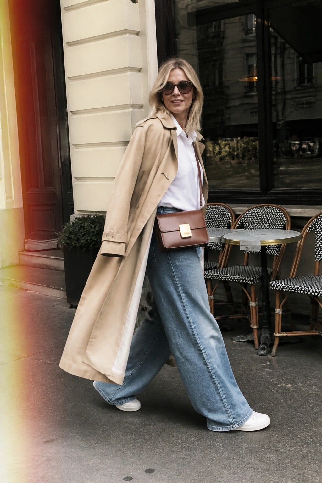 Blonde woman in beige trench coat and wide-leg jeans walking past outdoor cafe