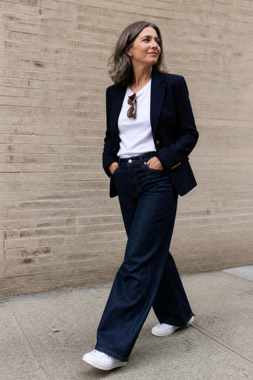 Woman over 50 in a navy blazer over white tee with dark wide leg jeans and white sneakers standing against a brick wall.