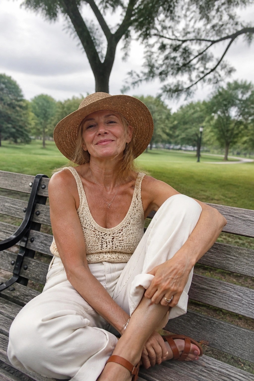 Woman in straw hat and crocheted top smiling on wooden park bench