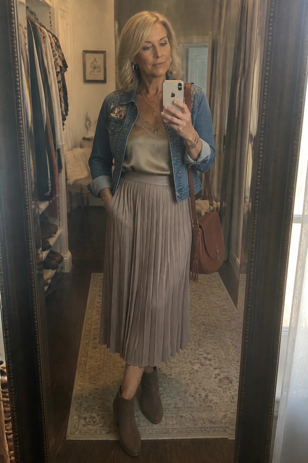 Blonde woman in embroidered denim jacket and pleated skirt taking mirror selfie with phone.