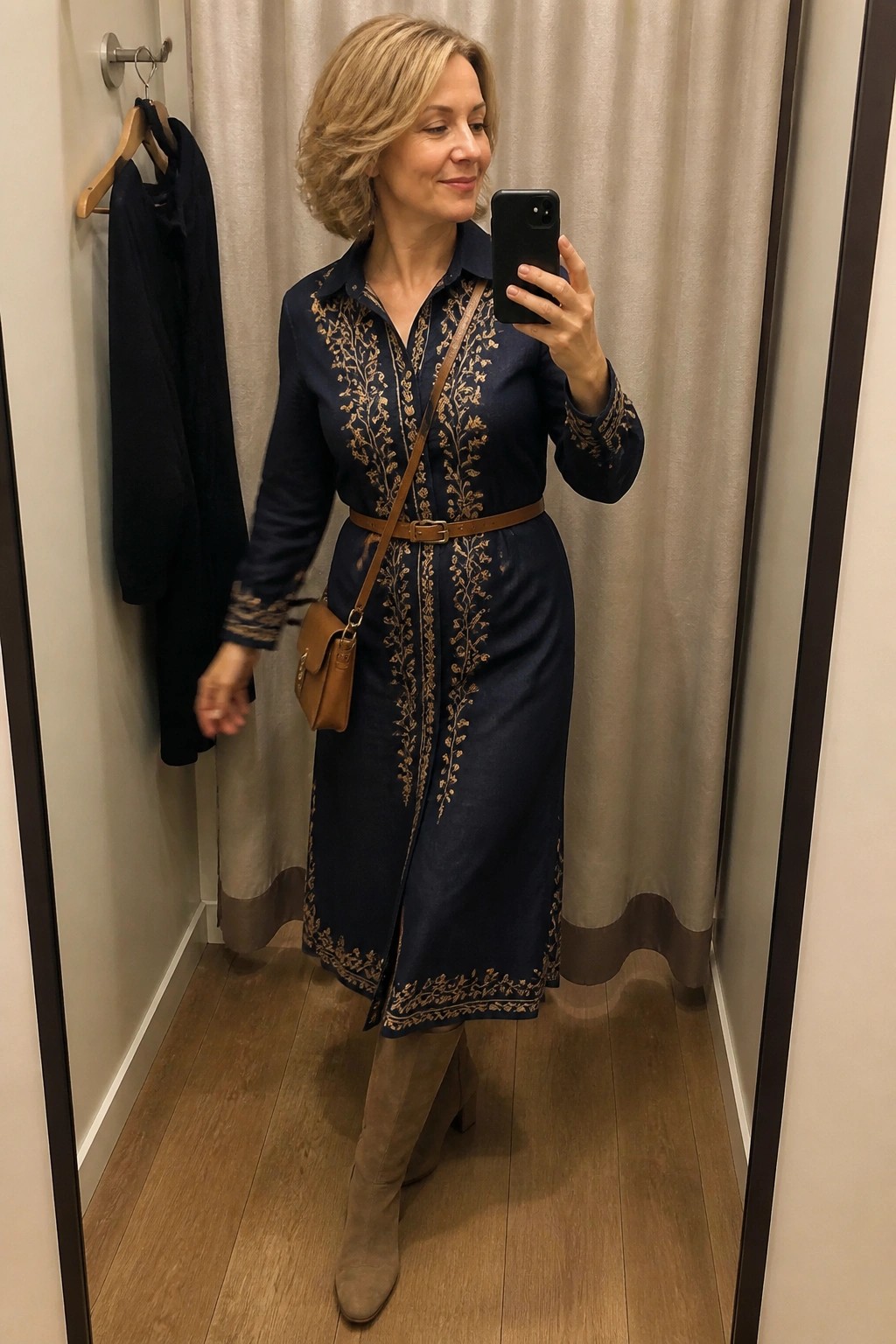 Woman in navy embroidered dress taking mirror selfie with brown bag and boots.