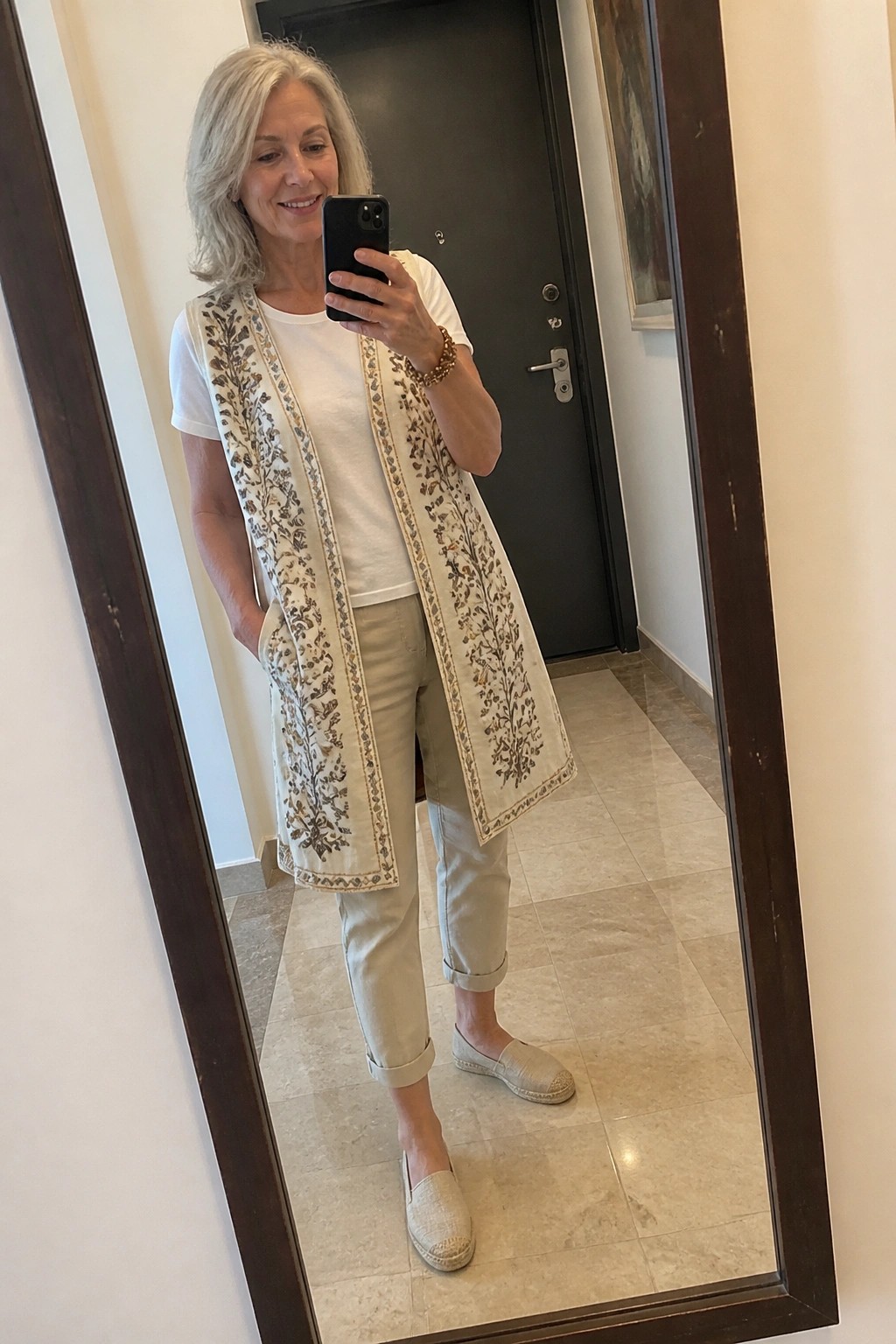Woman with gray hair taking mirror selfie in embroidered vest and beige pants.