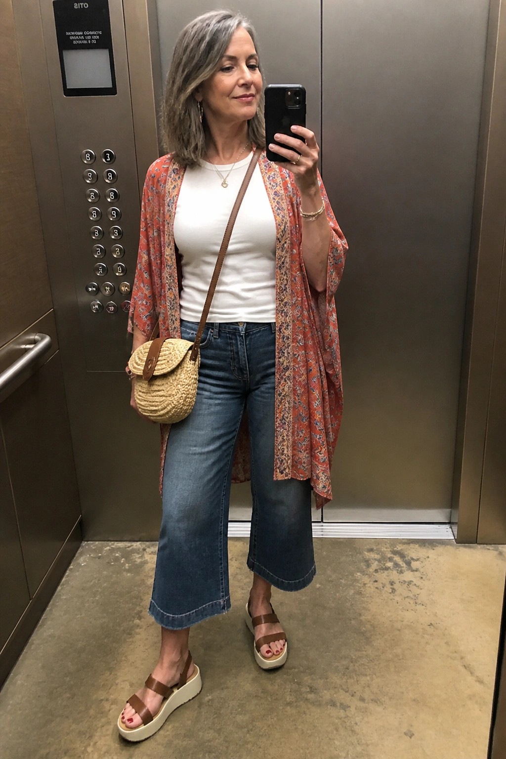 Woman in red patterned kimono, white top, and jeans taking elevator selfie with straw bag