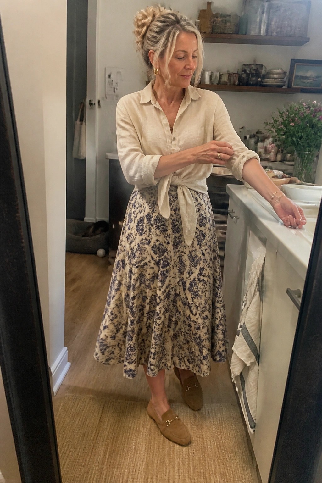 Blonde woman in tied cream shirt and floral skirt standing in kitchen