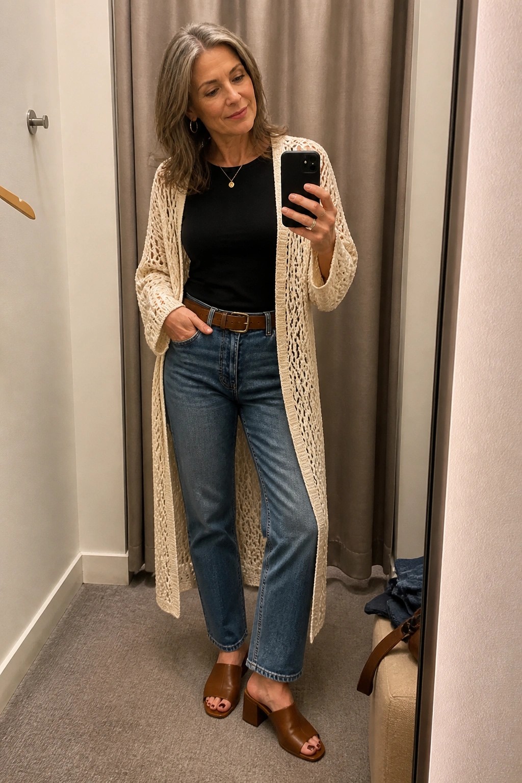 Gray-haired woman in cream cardigan, black top, jeans, taking mirror selfie.
