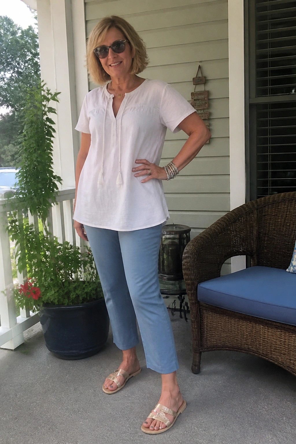 Woman in white blouse and blue pants posing on porch by wicker chair.