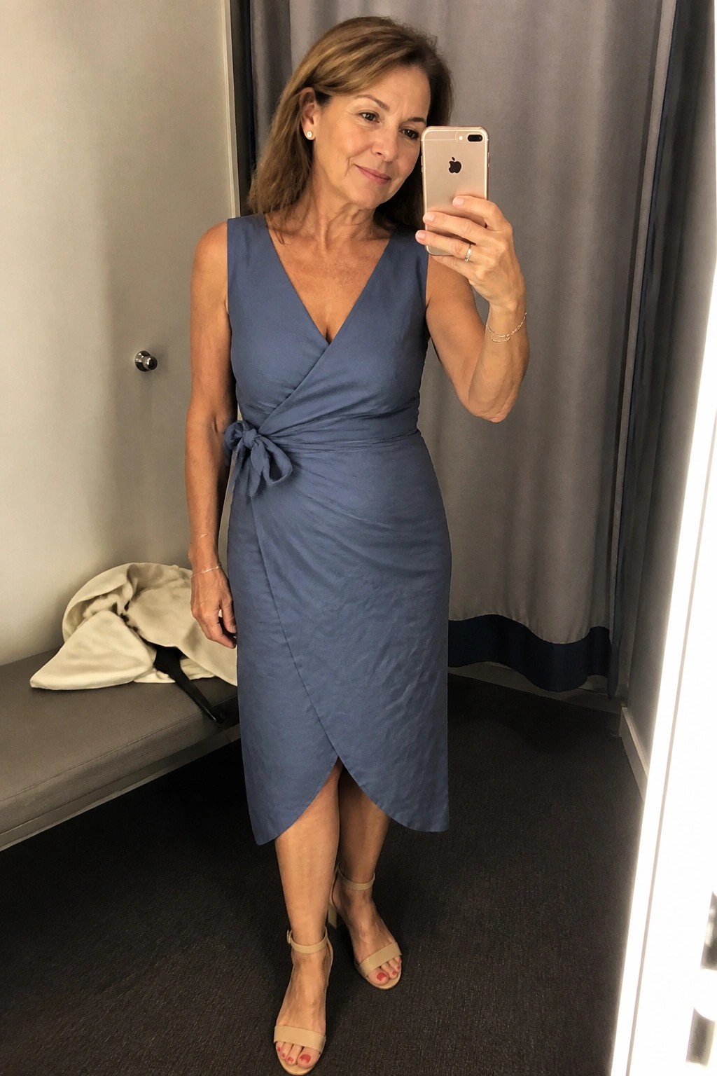 Woman in blue wrap dress taking mirror selfie in fitting room with phone.
