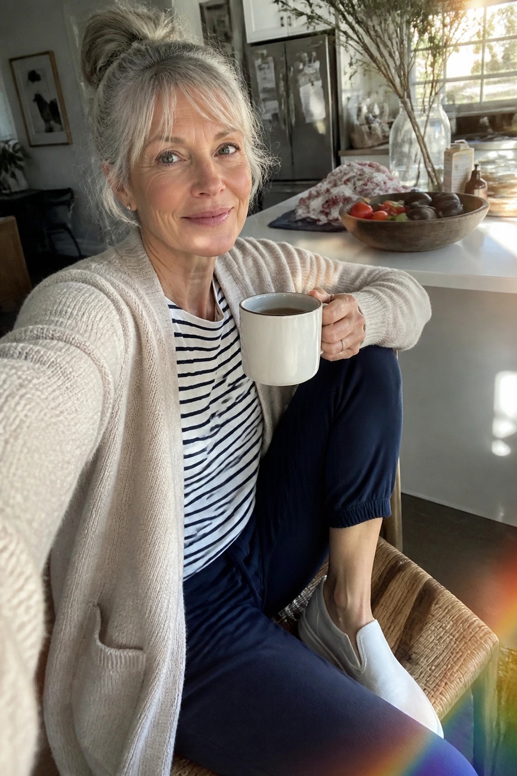 Woman with gray hair in bun wearing beige cardigan holds mug in sunny kitchen