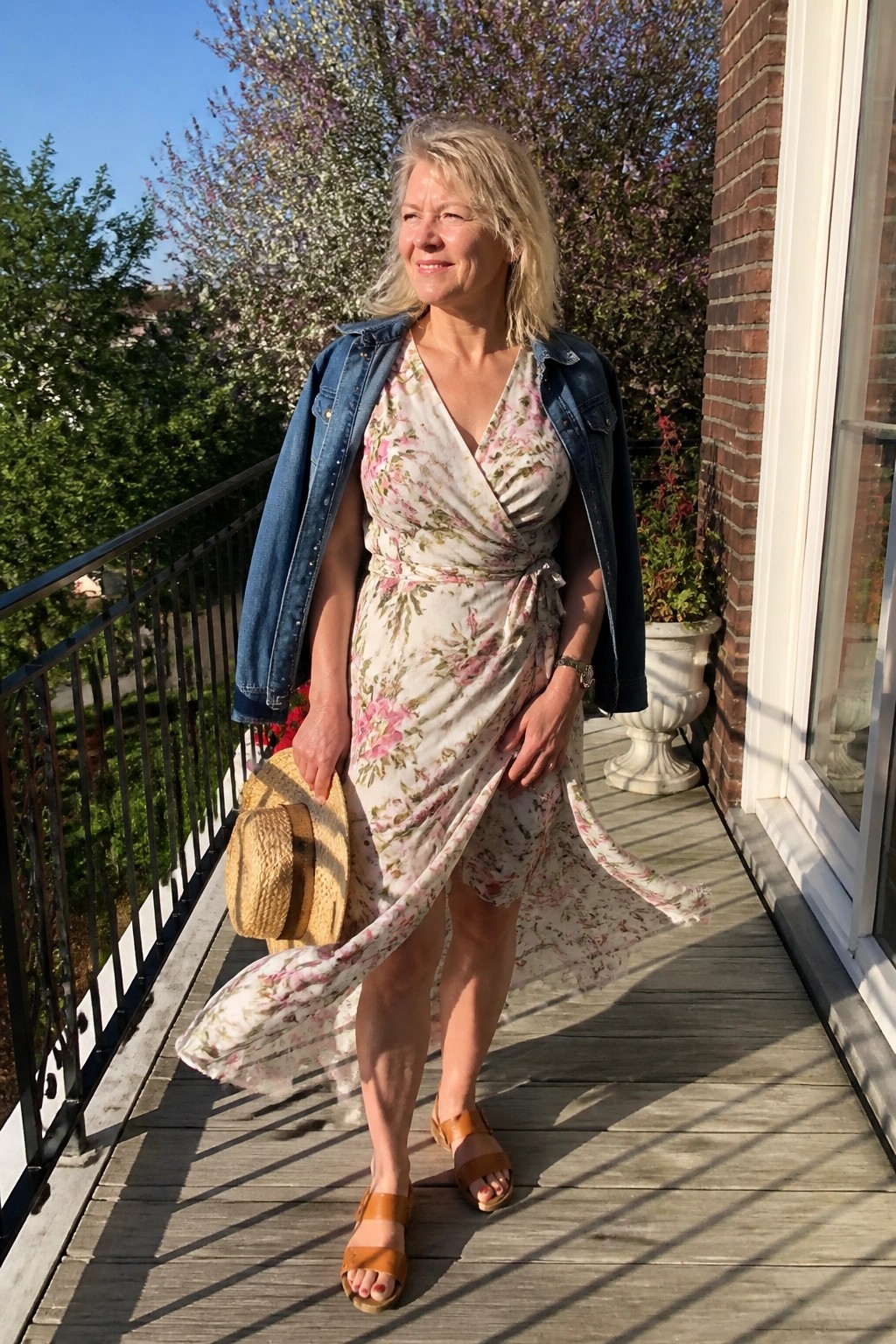 Woman in floral wrap dress and denim jacket holding straw hat on sunny balcony