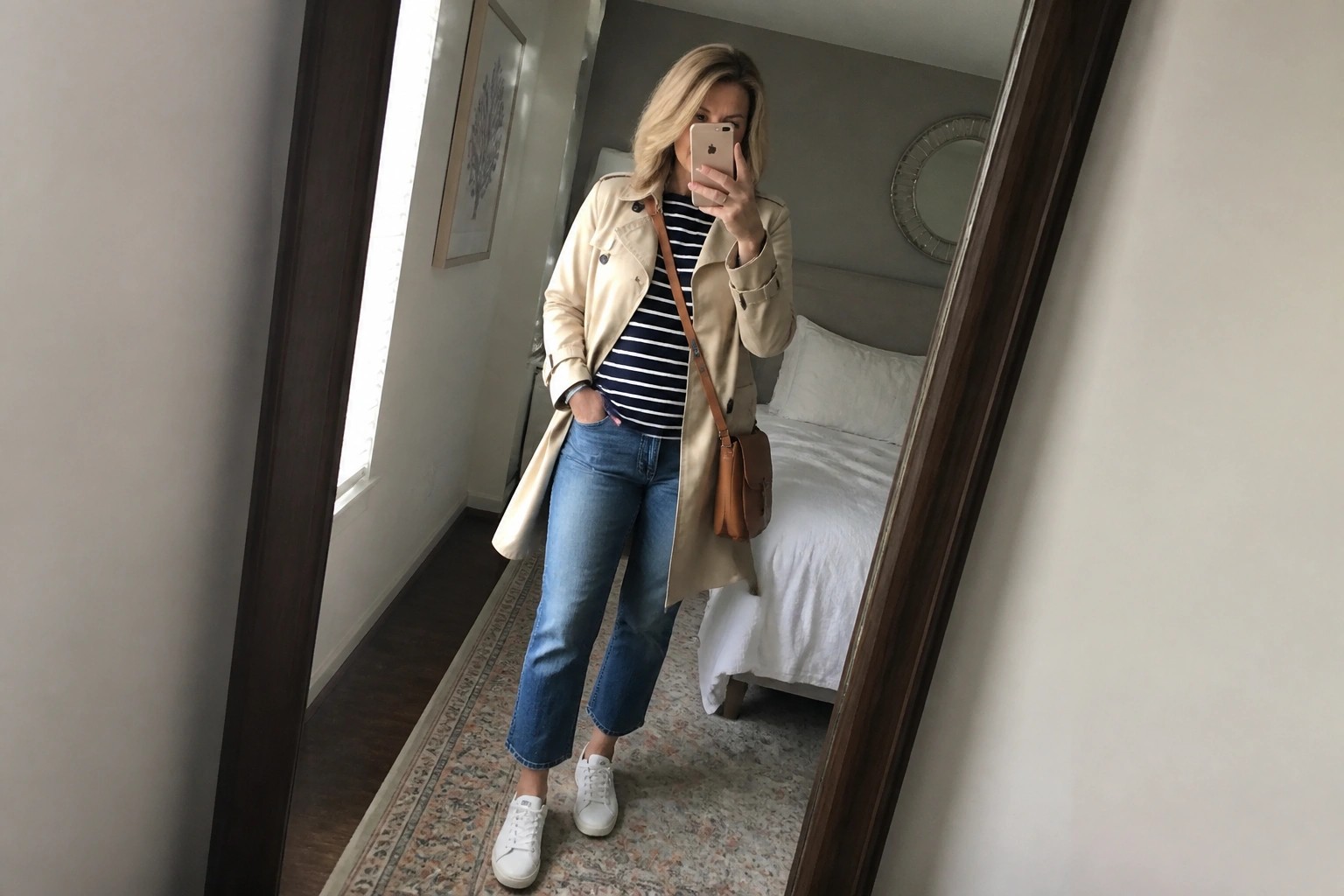 Woman in beige trench coat and striped shirt taking mirror selfie