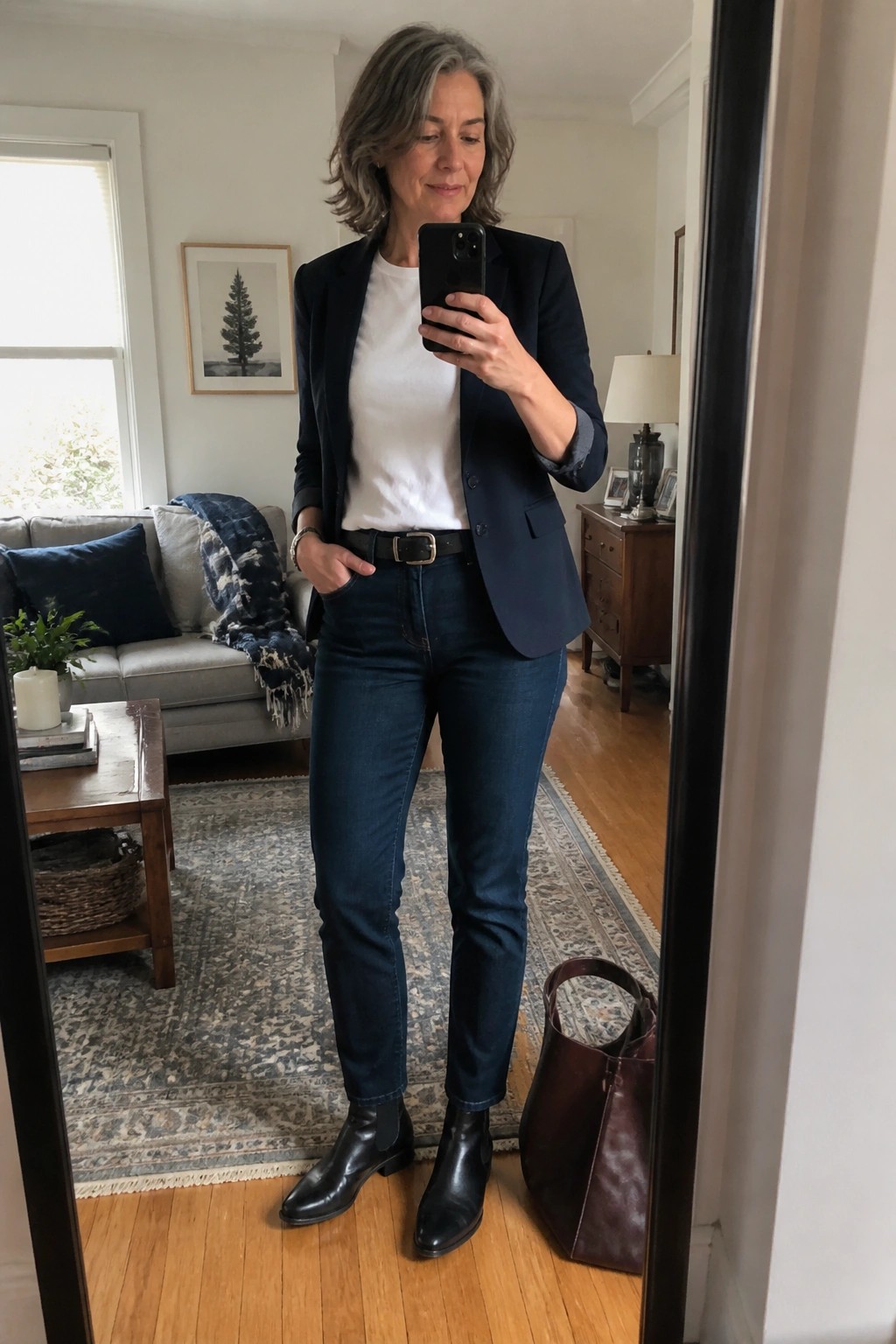 Gray-haired woman in navy blazer and jeans taking mirror selfie in living room