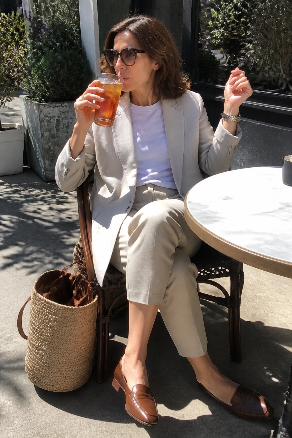 Woman in beige blazer sipping iced drink at sunny outdoor marble cafe table with woven bag