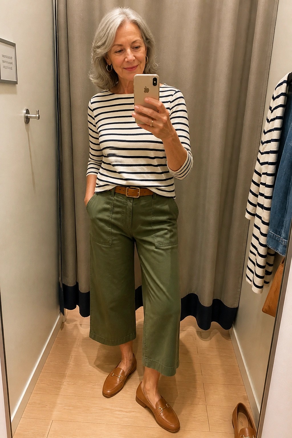 Silver-haired woman in striped shirt and green pants taking mirror selfie in fitting room.
