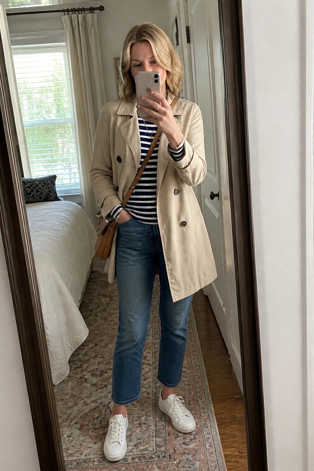 Woman in beige trench coat and striped shirt taking mirror selfie