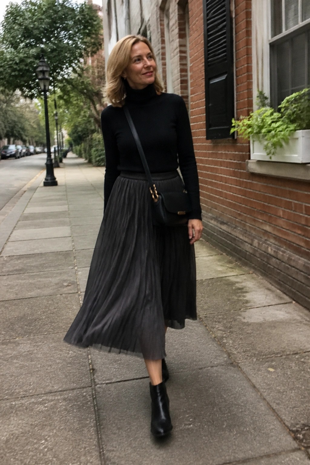 Woman in black turtleneck and pleated gray skirt with crossbody bag on sidewalk.