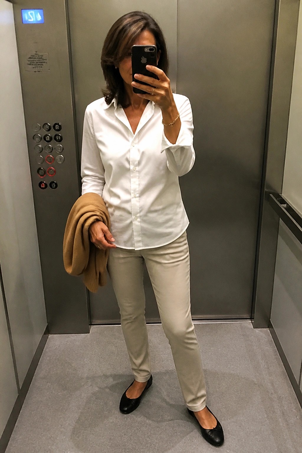 Woman in white shirt and beige pants taking selfie in elevator holding tan jacket