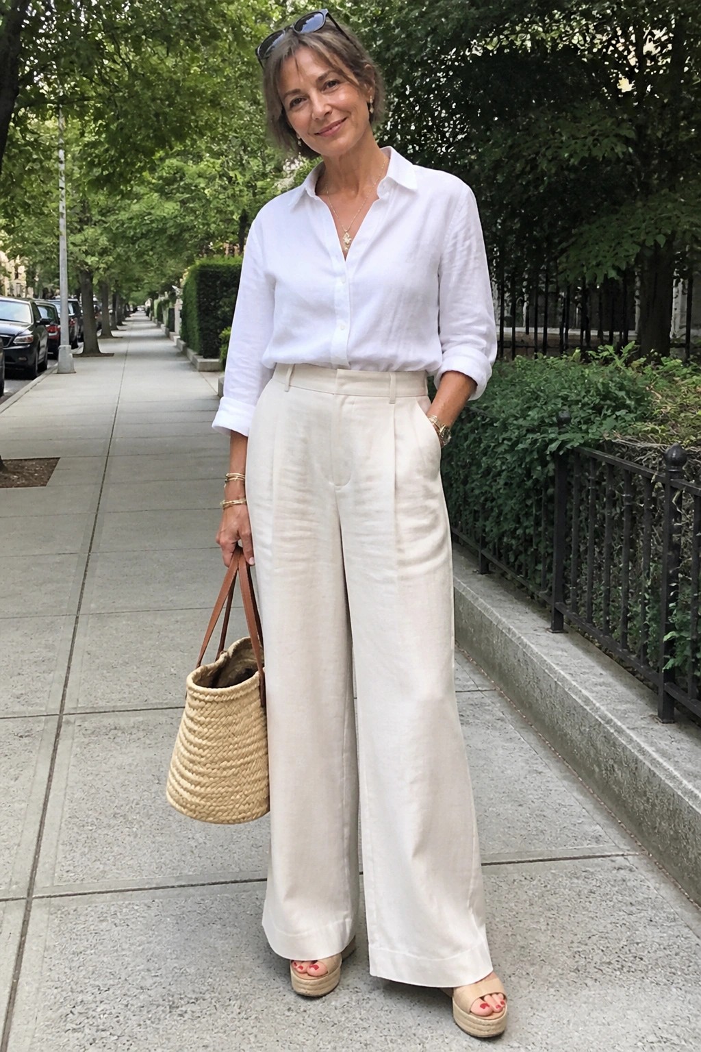 Woman in white shirt and cream wide-leg pants holding woven straw bag on sidewalk