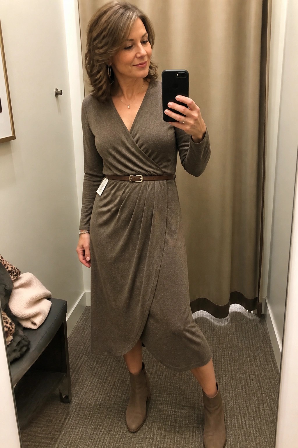 Woman in olive green wrap midi dress with brown belt and taupe ankle boots taking a mirror selfie in a fitting room.