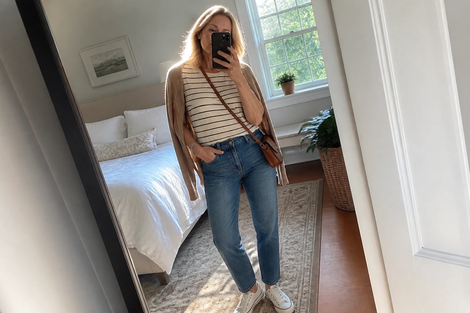 Woman in striped shirt and cardigan taking mirror selfie in bedroom wearing jeans and sneakers