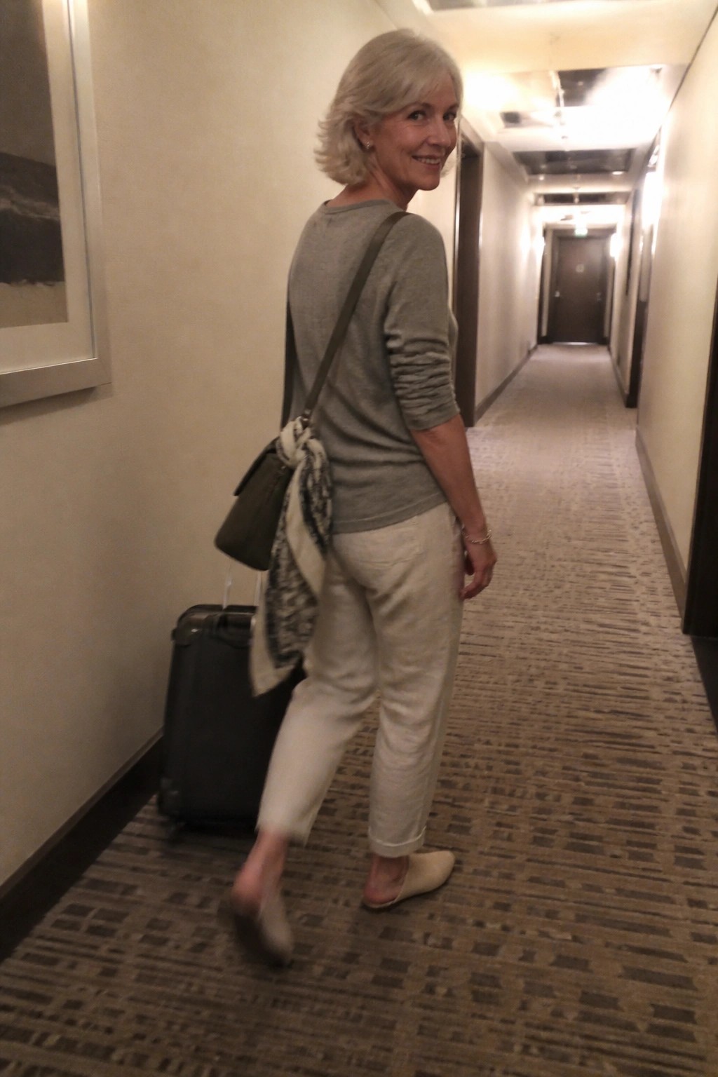 Gray-haired woman in gray top and white pants pulling black suitcase in hallway