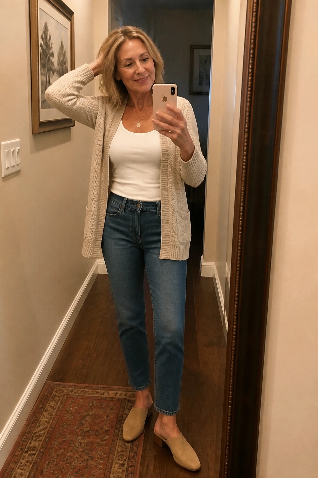Blonde woman in beige cardigan over white tank top and blue jeans takes mirror selfie.