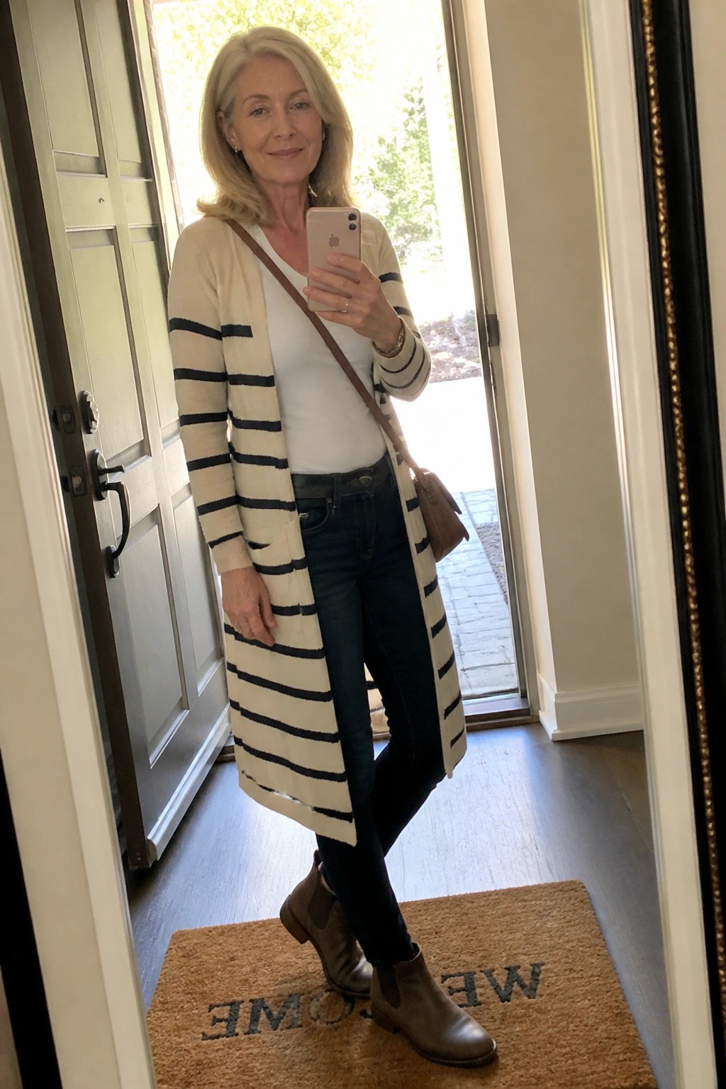 Woman in striped cardigan taking mirror selfie by welcome mat in entryway