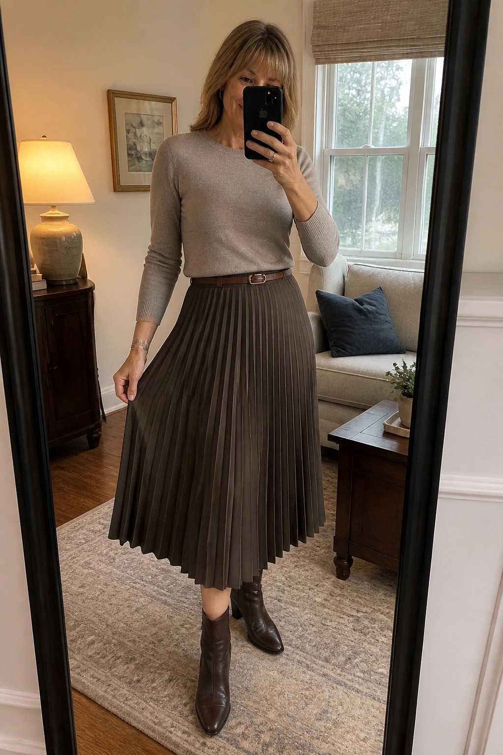 Woman in beige sweater and brown pleated skirt taking mirror selfie in living room