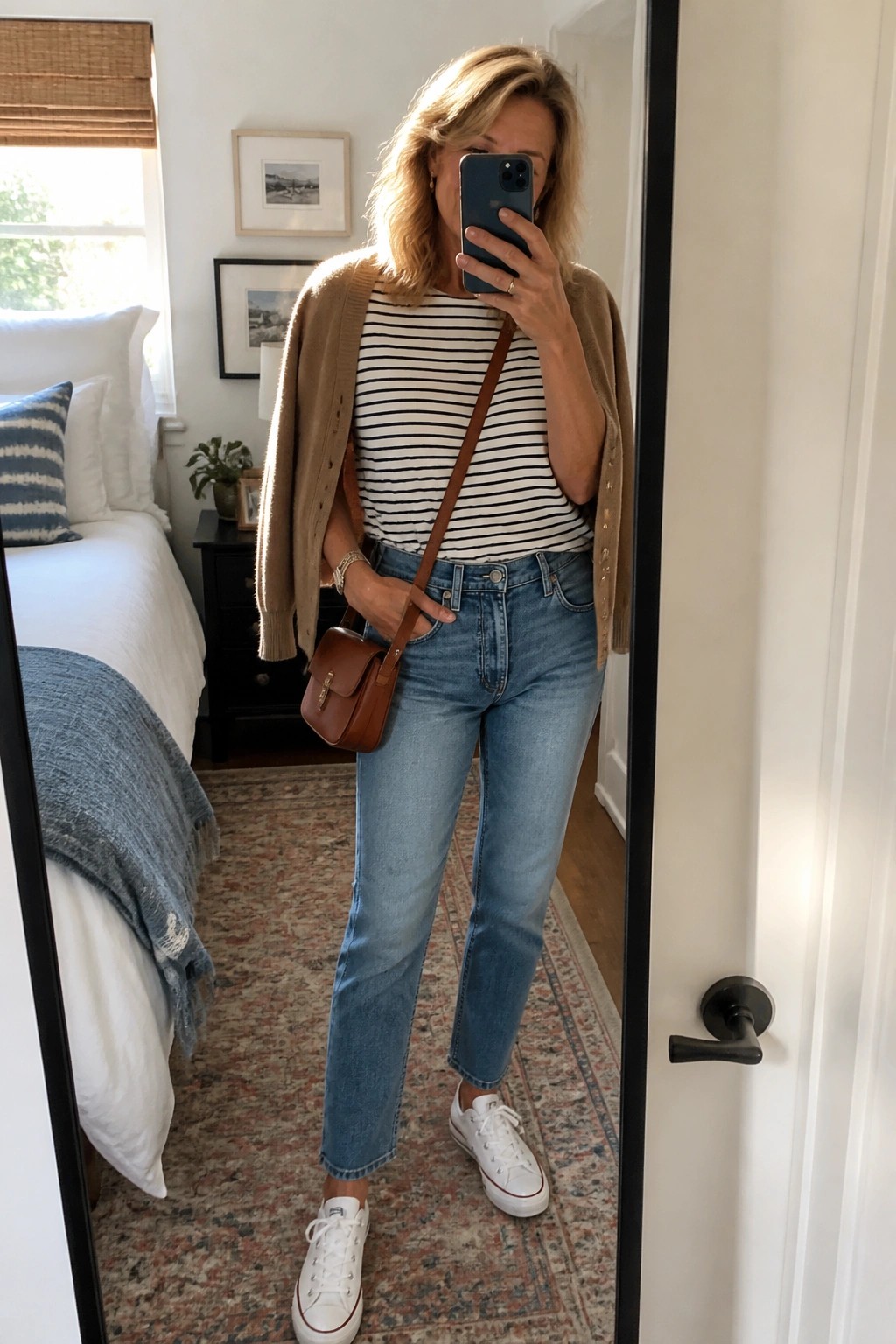 Woman in striped shirt and cardigan taking mirror selfie in bedroom wearing jeans and sneakers