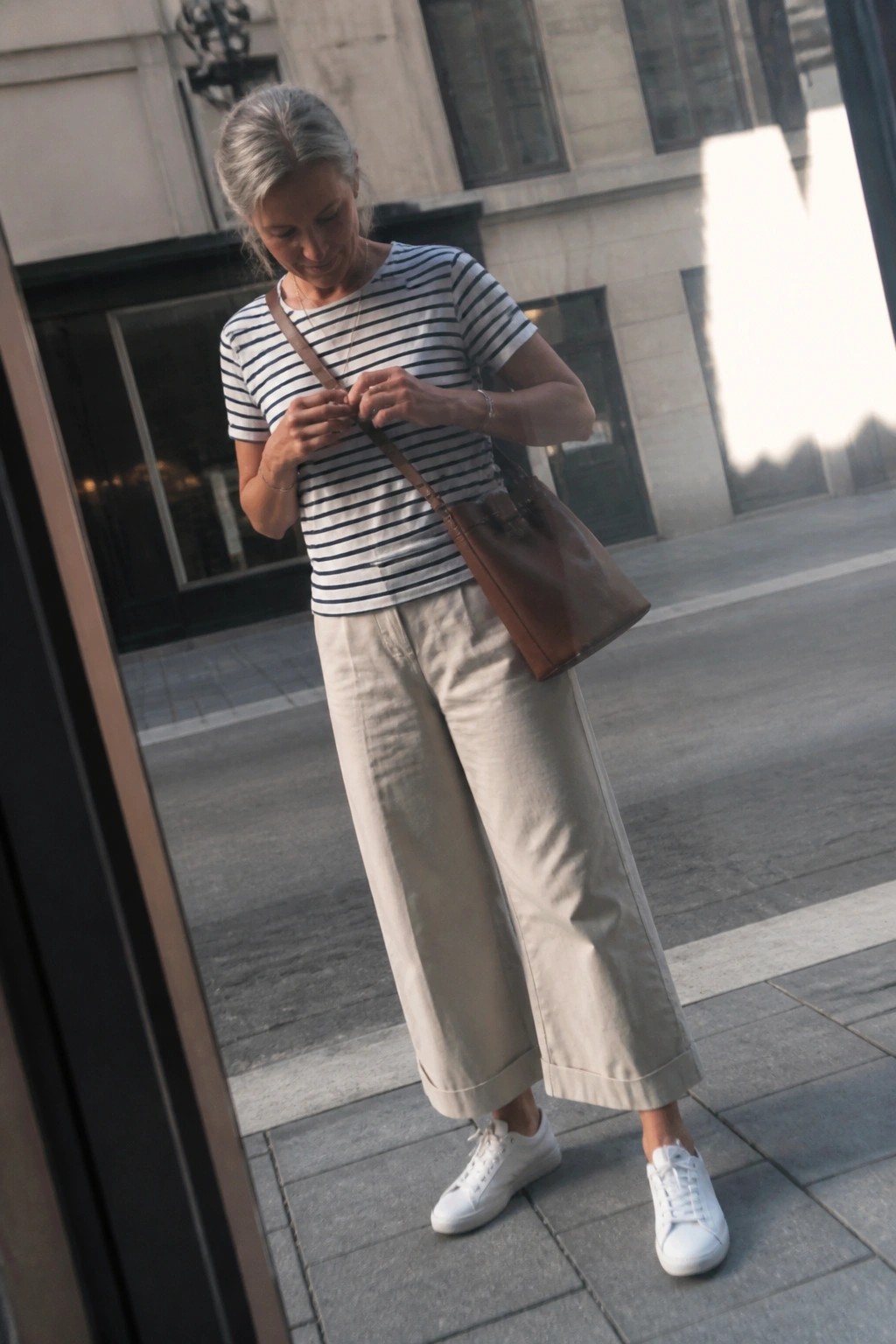 Gray-haired woman in striped shirt and beige wide-leg pants with brown leather bag.