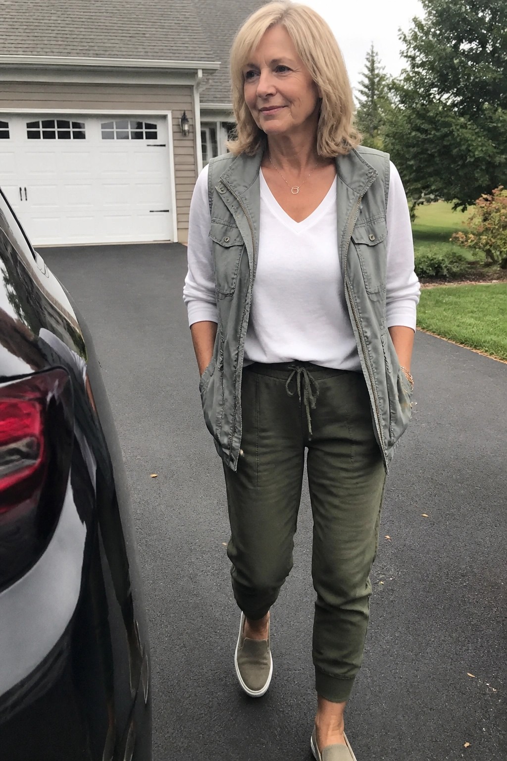 Woman over 50 wearing a sage green utility vest over a white long sleeve tee with olive green jogger pants and matching slip on sneakers.