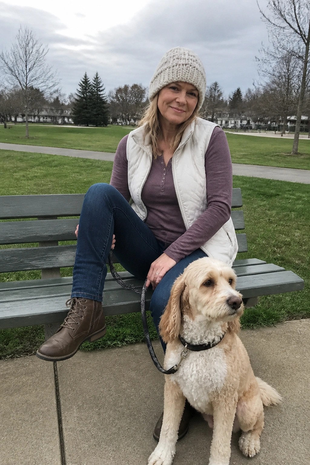 Woman in knit hat and white vest sits on park bench with beige dog