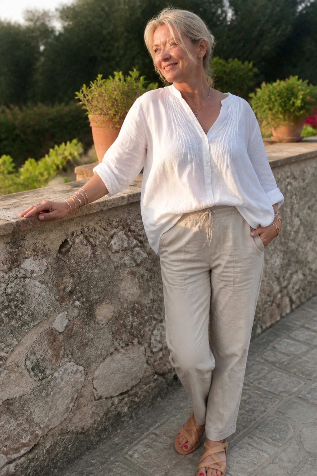 Smiling blonde woman in white blouse and beige pants leans on stone wall.