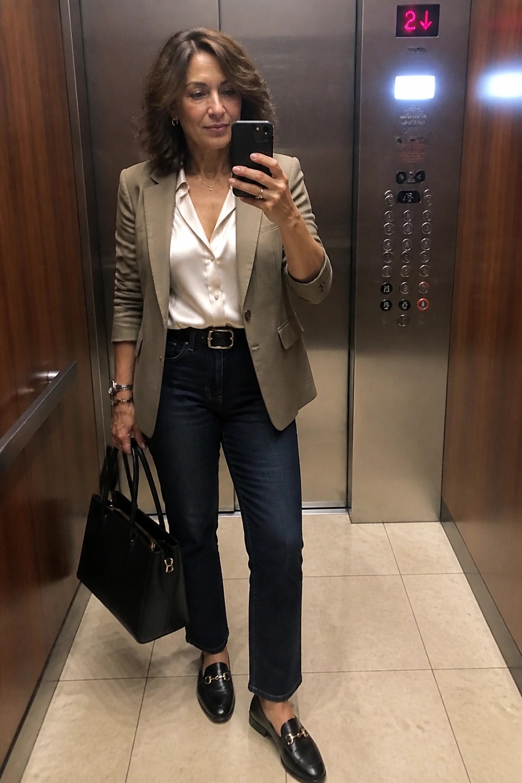 Woman in beige blazer and white shirt taking selfie in elevator with black bag