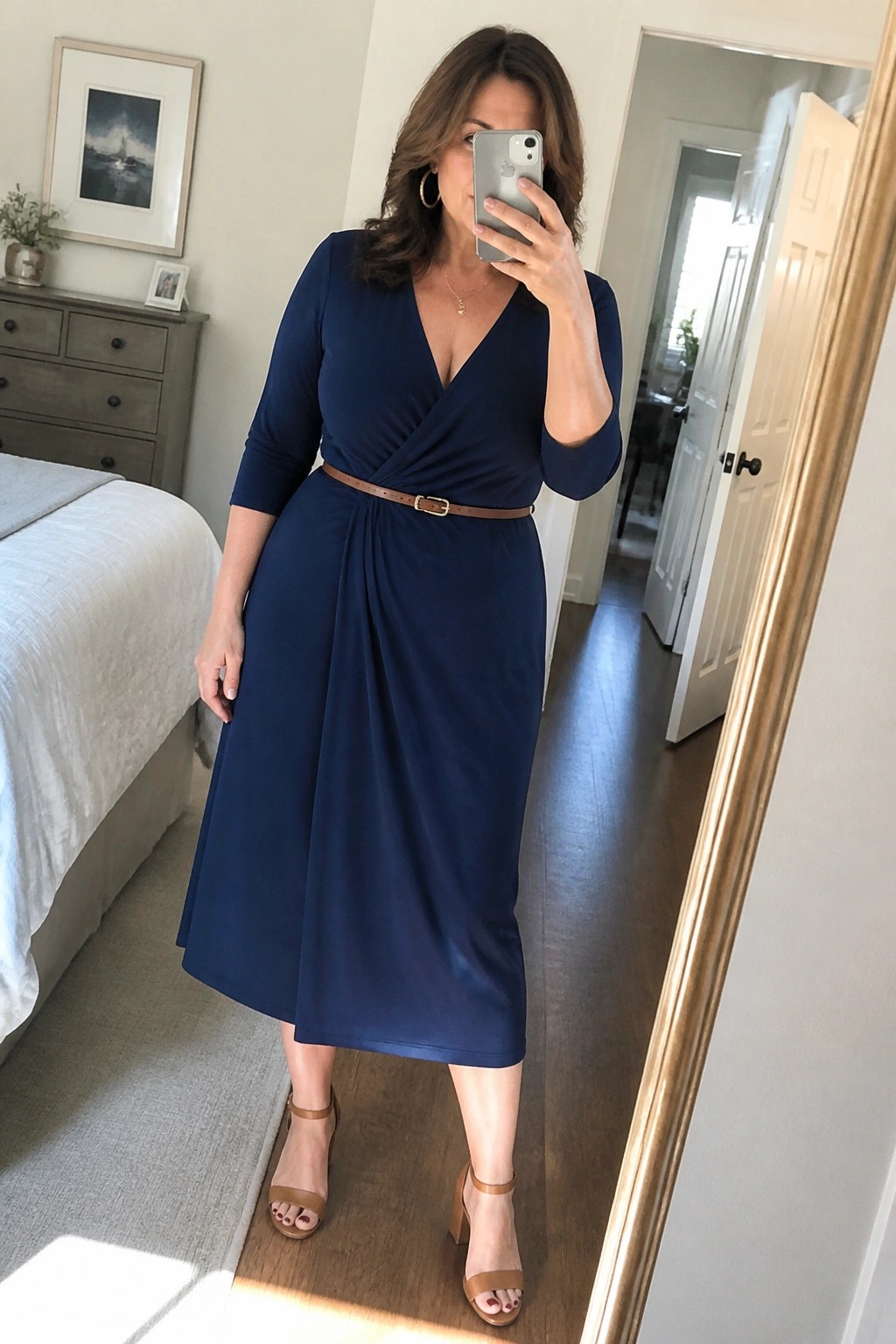 Woman in navy wrap dress taking mirror selfie with phone in bedroom