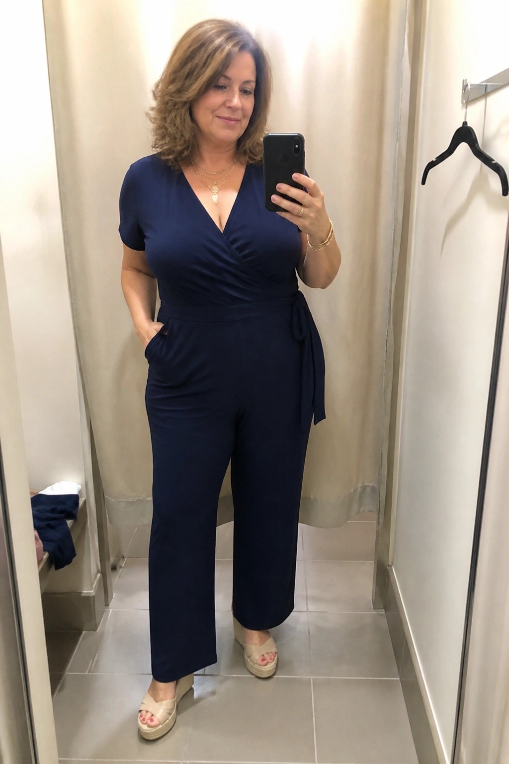 Woman wearing a navy blue short sleeve wrap jumpsuit with side tie and wide legs paired with beige wedge espadrilles and layered gold necklaces while taking a mirror selfie.