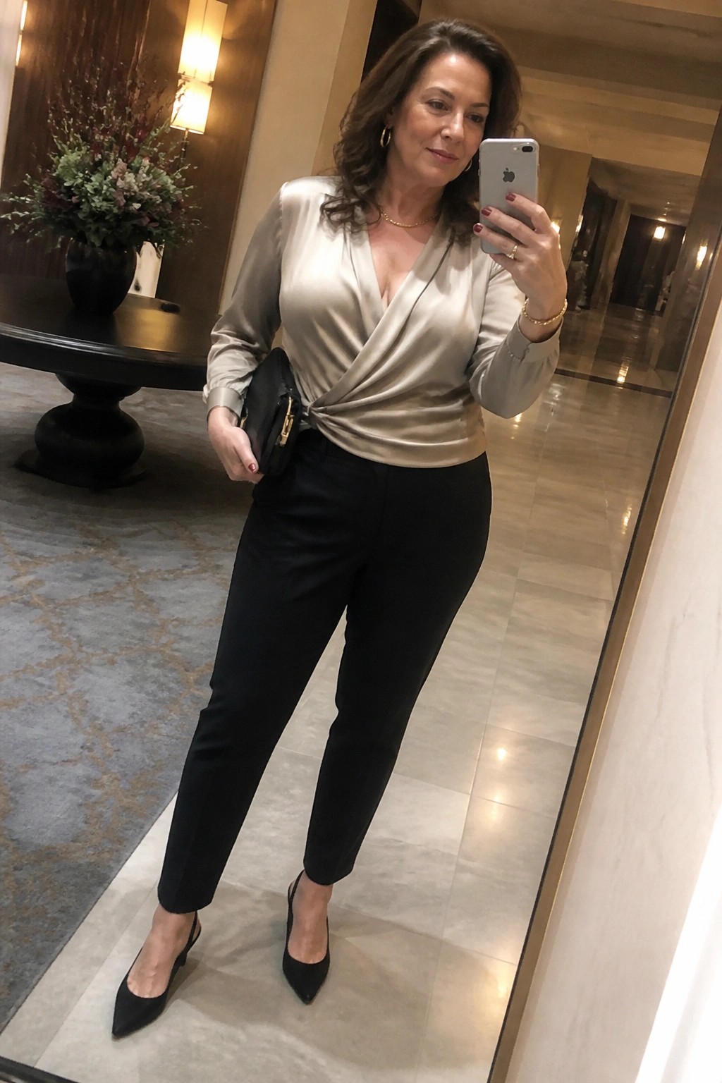 Woman in beige satin wrap blouse and black pants taking mirror selfie in hallway