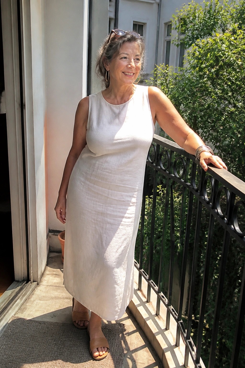 Woman in white sleeveless dress smiling on sunny balcony with hand on railing